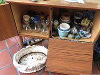 Wide view of two wooden cabinet shelves showing an assortment of plant pots, glass vases, ceramic jars and bowls in multiple sizes.