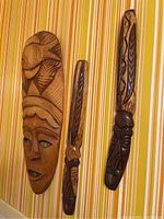 Three carved wooden items hung on striped yellow wallpaper, one large mask and two elongated figures.