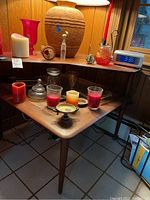 Full corner table showing tapered legs, cane top with various candles and glass items on it, wood paneling background, tiled floor.