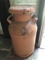 Full front view of a metal milk can showing surface rust, wear and dual side handles, photographed standing on floor near window.