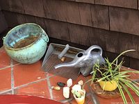 Shows large green ceramic pot, silver elephant watering can, small yellow pot with plant, decorative stones, and scooper on tile floor.