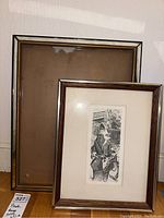 Both frames side by side leaning against wall: smaller wood-tone framed print in front of larger empty gold-tone frame