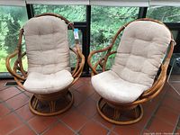 Pair of rattan rock and swivel chairs with beige tufted cushions in a sunroom setting.