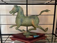 Full view of greenish metal galloping horse sculpture mounted on reddish wooden base, displayed on shelf