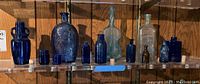 Shelf display of multiple vintage glass bottles in cobalt blue, light blue, clear, and brownish colors, varying shapes and sizes including decorative embossed designs and cork stoppers.