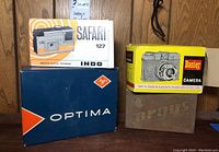 Four vintage cameras including Safari 127, Banier 120mm, Argus Model A and Agfa Optima boxes shown on wooden shelf