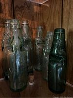 Seven vintage Codd neck glass bottles arranged on wood shelf, showing green and clear glass varieties with visible trapped marbles.