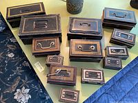 Thirteen vintage black and gold/copper tone metal boxes with assorted sizes and handle styles arranged on a table, showing general condition and variety.