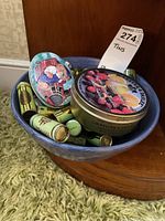 Photo of blue bowl containing multiple small vintage tins, including a round fruit-themed tin labeled McKeever & Dalzell, a small Santa Claus oval tin, and several cylindrical Tums tins.