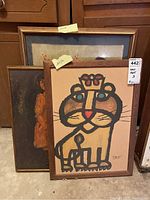 Three framed artworks leaning against cabinet: lion print front, portrait and botanical print behind
