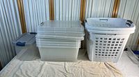 Stacked clear plastic storage containers with gray lids and blue latches, two white laundry hampers to the right