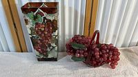 Front view of the stained glass vase with grapevine design next to red glass bowl with plastic grapes on cloth surface.