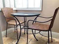 Bistro table with two matching rattan style chairs positioned near window showing full set from the side.