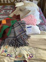 Multiple blankets and pillows piled on a quilted bed. Visible items include a plaid Pendleton blanket, a pink crochet blanket, a cherub embroidered blanket, and several decorative pillows.