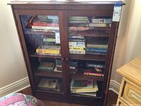 Front view of dark wood bookcase with glass doors showing books inside, two door knobs, rectangular shape, placed against a white wall.