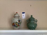 Two ceramic vases on shelf: flowered vase with lid and green vase with handles and lid