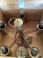 Top-down view of gold tone metal chandelier fixture in box showing five bulb sockets, attached chain, and bulbs.
