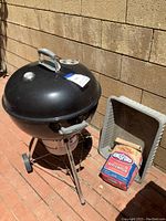 Weber charcoal grill and charcoal bag next to a gray plastic container outdoors.