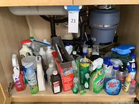 Multiple cleaning supplies inside a kitchen cabinet including various spray bottles, gallon jugs, steel wool box, and sponges