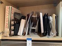 Various baking pans including metal muffin pans and a pizza stone stacked in a kitchen cabinet.