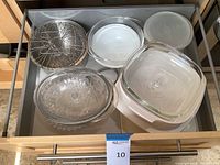 Drawer view showing multiple clear Pyrex glass dishes with lids, a flowered glass serving dish, round metal wire rack, and white storage container with lid.