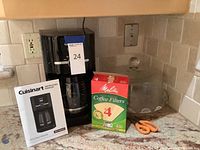 Black Cuisinart coffee maker setup on countertop with instruction manual, box of size 4 coffee filters, pair of orange-handled scissors, and clear glass cake platter with dome lid.