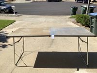 Full view of the folding metal table outdoors, showing one side raised and the other folded flat with metal tubular legs supporting it.