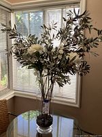 Tall clear glass vase with faux white flowers and olive branches, placed on a glass table near a window. The vase is filled with decorative stones and the faux plants rise about 47 inches tall.