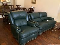 Two green leather armchairs with rolled arms and nailhead trim, plush cushions, and wood legs, situated on wood flooring beneath wall art and near dining furniture.