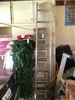 Three aluminum ladders leaning in a garage. Visible are a short stepstool ladder, a medium height ladder, and a taller ladder further back.