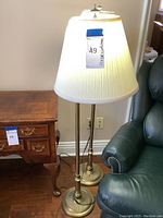 Two matching gold tone floor lamps with white pleated lamp shades next to a wooden table and armchair.