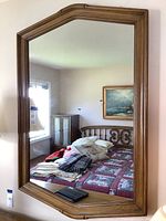 Full view of the large wooden framed mirror reflecting a bedroom interior with a bed, pillows, and wall art.