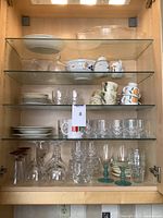 Glass cabinet shelf with assorted Pyrex baking dishes, stemware including wine glasses and Courvoisier glasses, Mikasa tea cups and plates with floral patterns, bowls and drinking glasses.