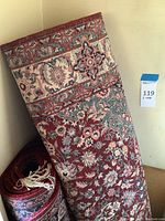 Rolled burgundy floral area rugs standing against wall, showing patterns and wear