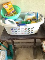 Dark wood side table with heavily used surface, white plastic laundry basket filled with laundry and cleaning products, and a green plastic basin inside the basket.