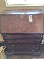 Front view of National Mt. Airy writing desk showing the dark stained wood slant front drop lid with brass lock and four drawers below with brass handles.