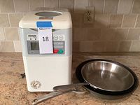 Zojirushi bread maker unit with two stainless steel frying pans stacked beside it on countertop.