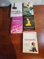 Six Douglas Coupland paperback novels on a wooden table, titles visible including The Gum Thief, Girlfriend in a Coma, Miss Wyoming, Hey Nostradamus!, Microserfs, and one unidentified title.