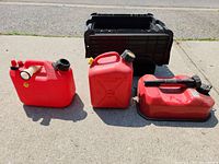 Three small red gas cans arranged on pavement outside with a black plastic crate behind them.