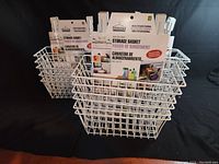 Photo showing white metal over-the-door storage baskets with hooks arranged in stacks, with packaging showing Maurice brand and storage basket label.