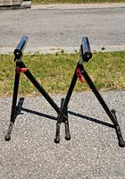 Two adjustable folding rolling stands with chrome roller tops and red adjustment levers standing on asphalt surface.