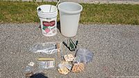 Assortment of wine making supplies spread on ground: white and translucent plastic fermentation buckets, multiple bags of corks, hand corker, tubes with pinch clamps, brush, and labels visible