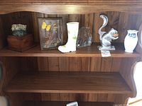 Wide view of wooden shelf holding Mats Jonasson art glass, Aynsley boot vase, Belleek vase, squirrel nutcracker, framed botanical display, and wooden box with stone.