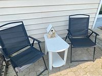 Two black folding chairs with mesh seats and backs flanking a small white resin plastic table with a birdhouse on top, all set outside on a paved surface near a beige wall.