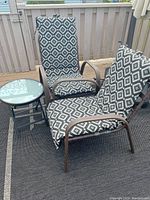 Two metal framed patio chairs with black and white diamond patterned cushions and a small round folding glass top patio table on a deck.