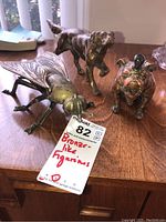 Three bronze-like metal figurines on a wooden surface: one large insect and two dogs, showing wear.