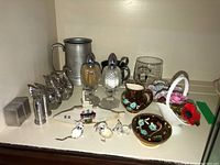 Overview of lot showing glass salt and pepper shakers, silver plated swan figurine, pewter mug, ceramic bowl set and basket with flower decoration, souvenir shot glass, small figurines and metal salt shakers.