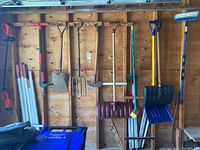 Wide view showing all shovels, stakes, pitchfork, mallet, Simoniz brush, and broom hanging on a wooden wall.