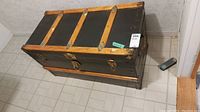 Diagonal front-top view of black travel trunk showing wooden slats and metal corner brackets.