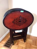 Photo of a wood TV table showing a dark wood oval folding top with an intricate black decorative design. The table is standing upright on the floor against a wall.